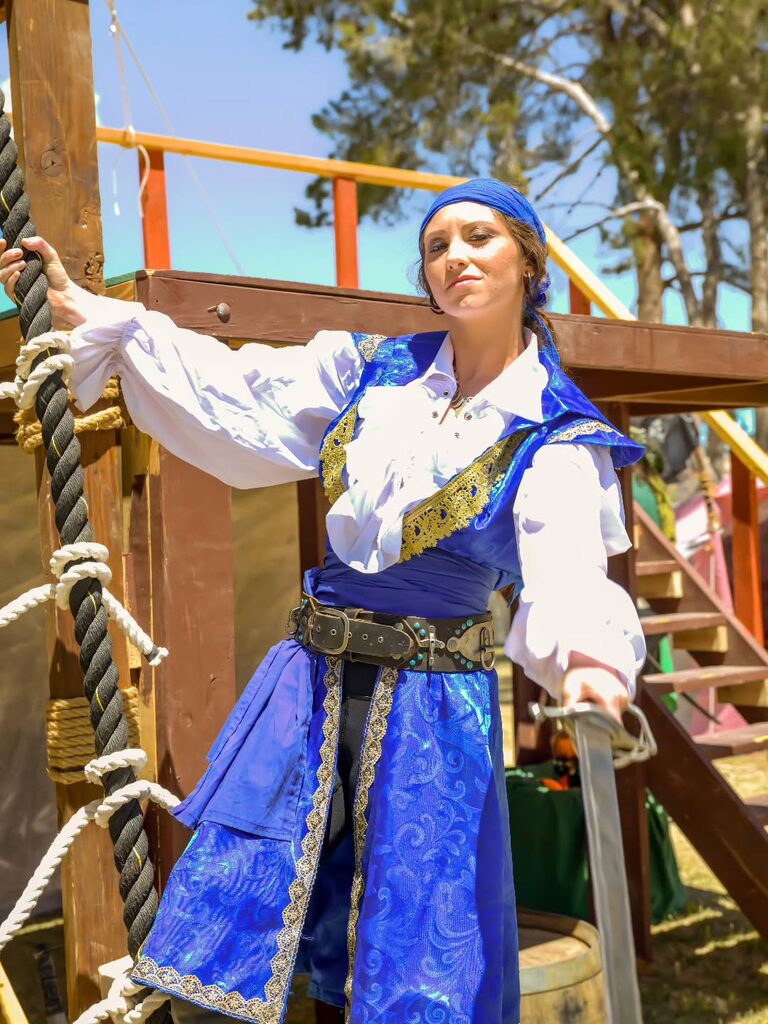 Woman in a blue pirate costume holding a rope and cutlass on a ship set.