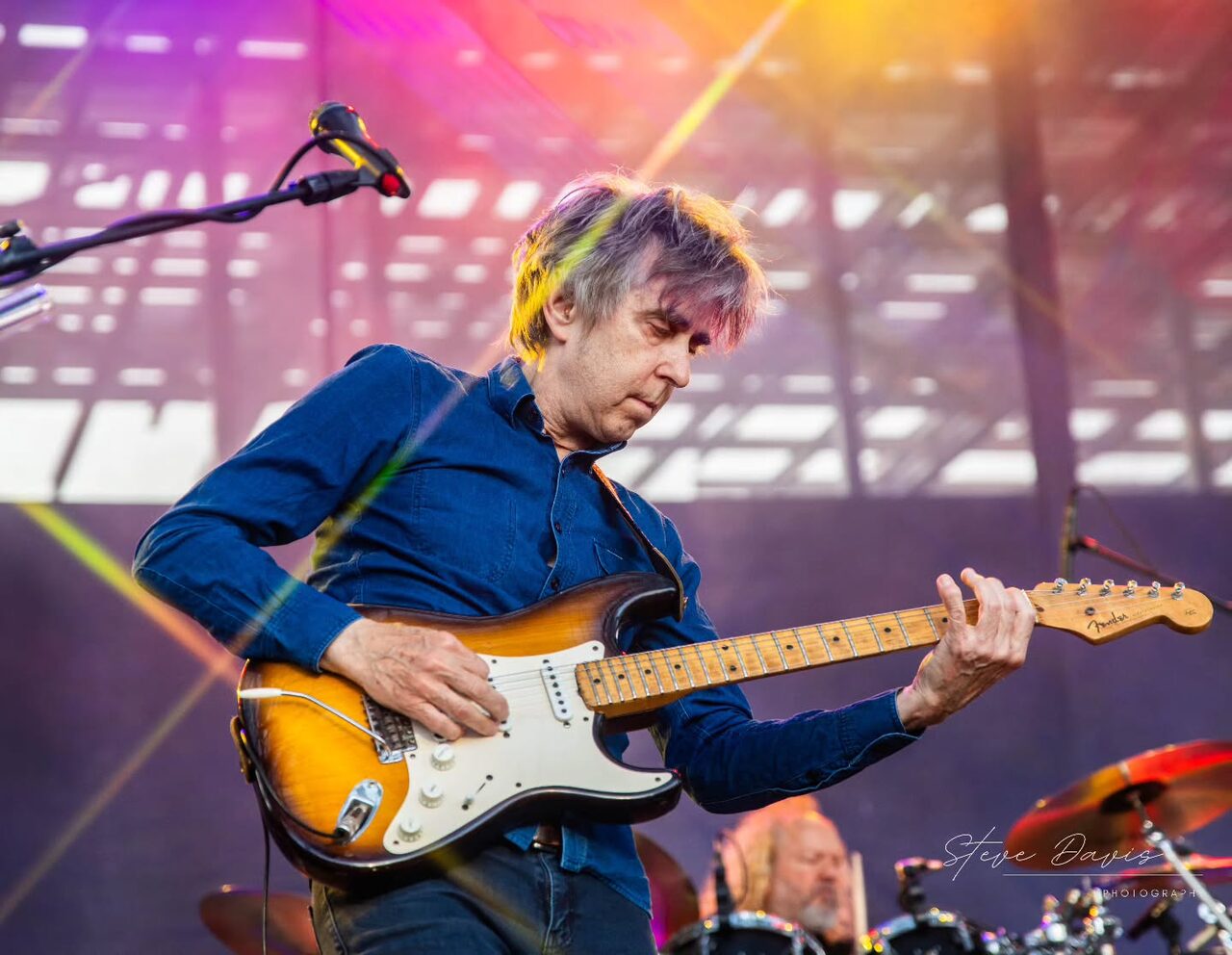 Eric Johnson playing a Fender Stratocaster beneath pink and yellow stage lights.