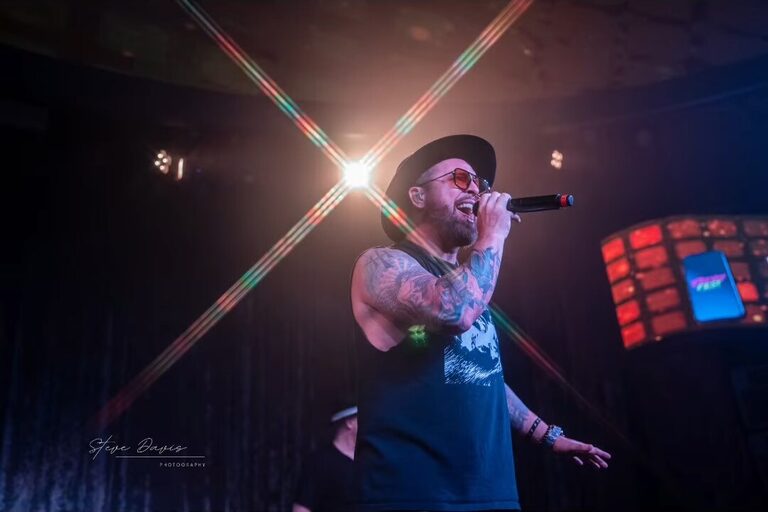 Tattooed singer holding a microphone beneath crossing stage lights.